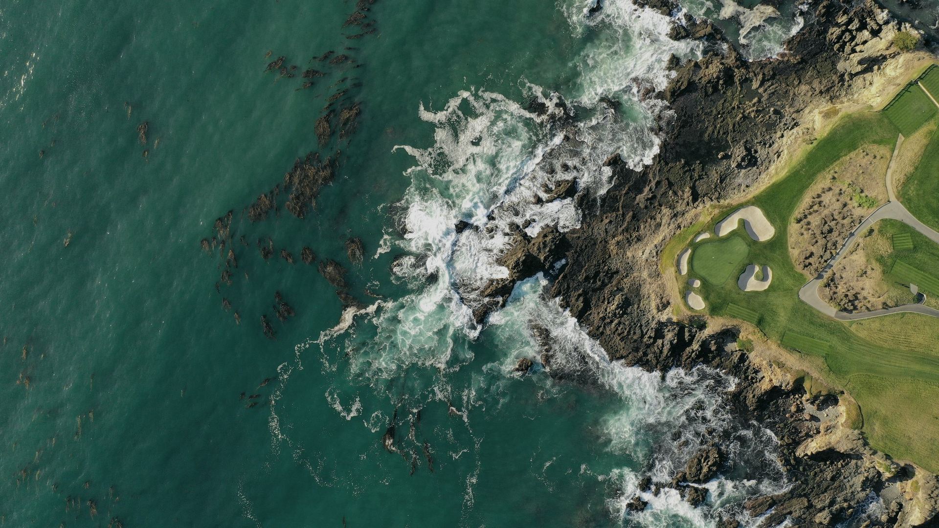 Aerial view of a coastal golf green ringed by rocks and breaking surf